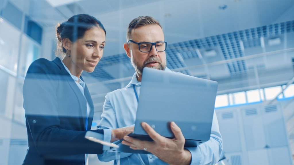 Stylish Confident Businesswoman and Businessman Working on a Project, Man Holds Laptop Computer, They Reference it. In the Background Modern and Bright Facility. Low Angle Shot