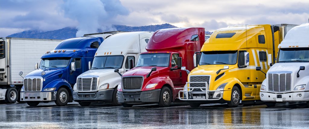 Different make big rigs semi trucks tractors with loaded semi trailers standing in the row on truck stop parking lot at early morning waiting for the route continuation time according to the log book