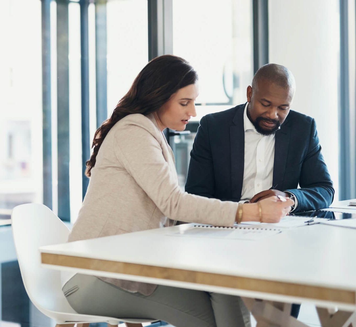 Businessman and business woman sitting at a desk discussing the specifics of a deal on a notebook.