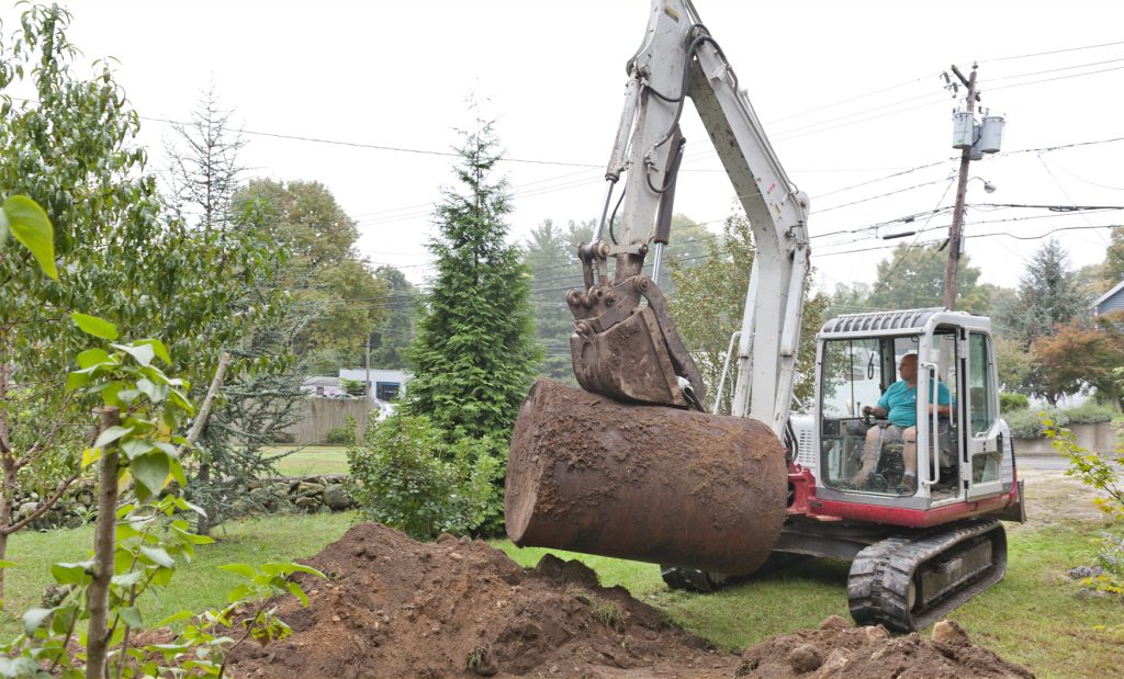 "A man sitting in a compact excavator removes an old, rusty underground oil tank from a residential yard."