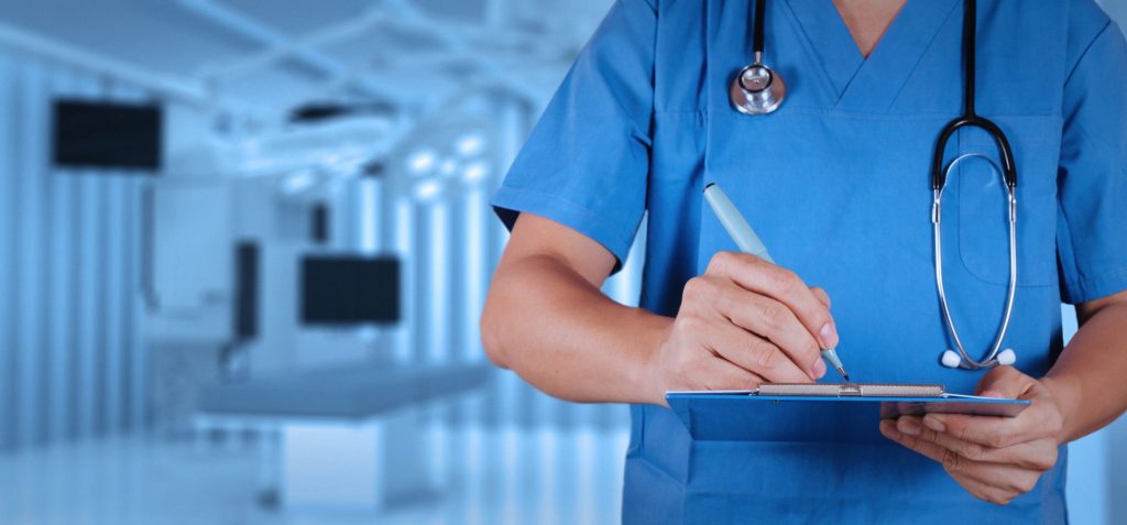 Medical provider in scrubs with stethoscope jotting down notes in an examination room